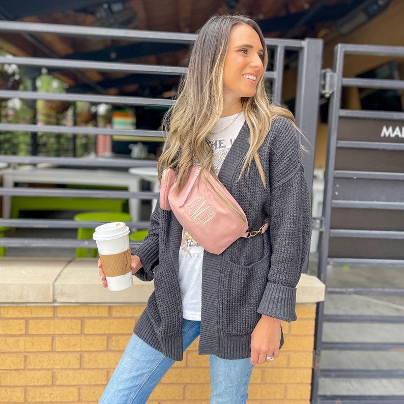 Blush colored, nylon sling bag across the chest of a model holding coffee. Model is also wearing a t-shirt and cardigan. Monogrammed letters in Ivory thread decorate the front of the crossbody. A webbed, adjustable strap is the same hue as the bag.
