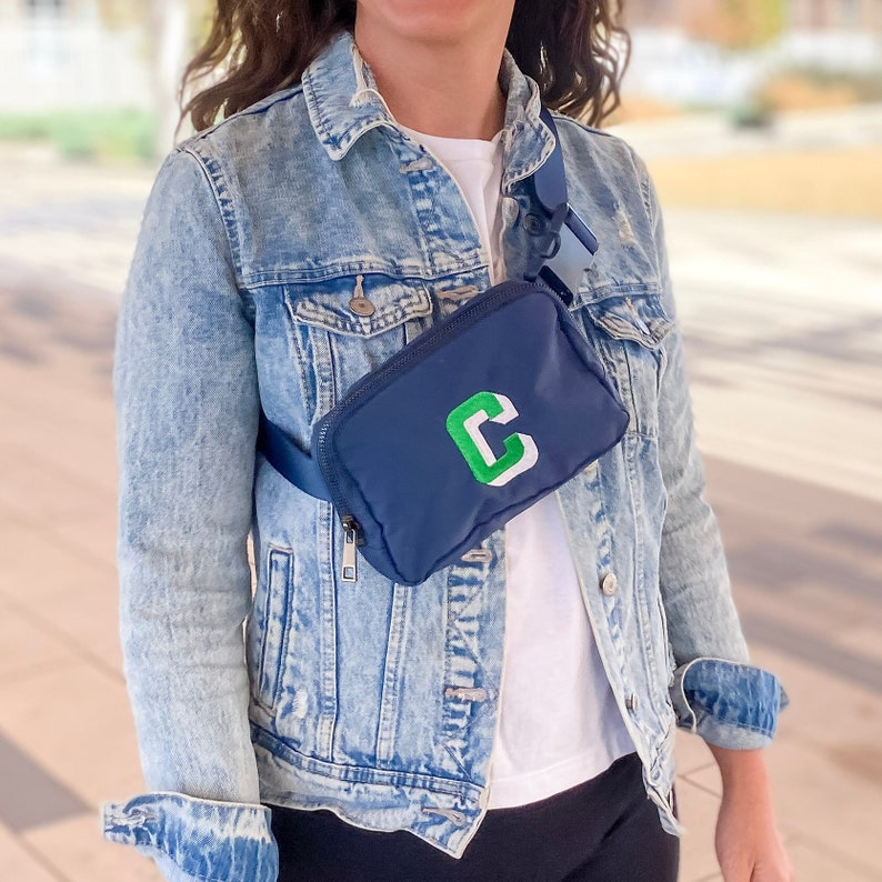 Nylon Navy Belt bag, worn across the chest of a model in a denim jacket. Fanny pack is embroidered with a Kelly green block "C" with a white shadow.