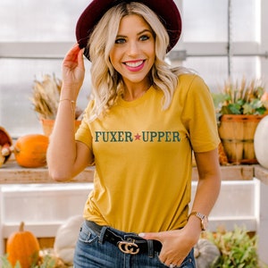 May include: A woman wearing a mustard yellow t-shirt with the text "FUXER * UPPER" in green and red. She is wearing a burgundy fedora hat and blue jeans. She is standing in a greenhouse with pumpkins and plants in the background.