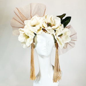 Könnte beinhalten: Ein weißer Mannequinkopf ist mit einem floralen Kopfschmuck geschmückt. Der Kopfschmuck besteht aus großen, cremeweißen Magnolienblüten, grünen Blättern und goldenen Quasten. Eine tanfarbene Netzfächerform und goldene Verzierungen vervollständigen das Design.