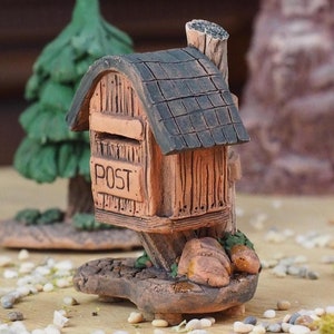 Op de afbeelding: Een miniatuur houten brievenbus met een bruin dak en het woord "POST" aan de voorkant, omringd door kleine groene bomen en witte kiezelsteentjes. Op de voorgrond staat een kleine witte kaars.