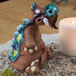 Könnte beinhalten: Eine Keramik-Drachenfigur mit grünen, blauen und gelben Akzenten sitzt auf einem weißen Keramiktablett mit kleinen Steinen und grünem Moos. Eine weiße Kerze und ein kleines Teelicht befinden sich ebenfalls auf dem Tablett.