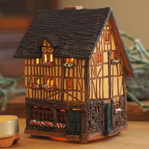 Op de afbeelding: Een klein, bruin en geel keramiek huis met een zwart dak en verlichte ramen. Het huis staat op een houten tafel met een kleine, witte kaars ervoor.