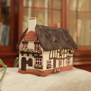Può includere: Un piccolo cottage in ceramica bianca con tetto di paglia e un cartello che recita "Old Trailor Tavern". Il cottage è decorato con fiori e ha finestre con luci accese. Il cottage è posto su una superficie di tessuto beige.