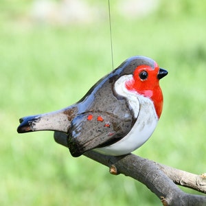 May include: A ceramic bird figurine with a red breast, gray wings, and a white belly, perched on a small wooden branch. The bird is suspended by a thin string against a blurred green background.