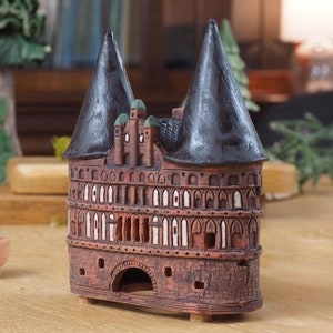 May include: A small, brown ceramic model of a medieval castle with black roofs and white accents. The model is sitting on a wooden table.