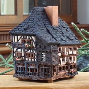 Può includere: Una piccola casa in ceramica nera e marrone dettagliata con un tetto nero e modanature bianche. La casa ha più finestre e un camino. La casa è appoggiata su una superficie di legno.