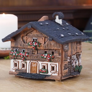 Könnte beinhalten: Ein Keramik-Räucherofen in Form eines kleinen, braunen Chalets mit schwarzem Dach und weißen Verzierungen. Das Chalet hat rote Blumen auf den Balkonen und einen Schornstein. Der Räucherofen steht auf einem Holztisch.