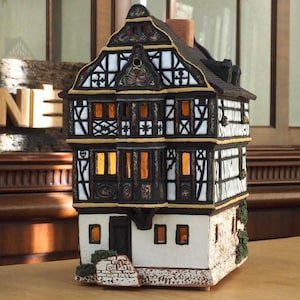 May include: A black and white ceramic house figurine with a half-timbered design and lit windows. The house is sitting on a wooden table with a wooden sign that reads "MIDENE" in the background.