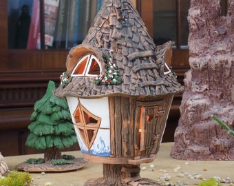 May include: A ceramic fairy house with a textured, brown shingle roof and a white and brown facade. The house has a small, open door and windows with a warm glow. Decorative trees and rock formations surround the house. The scene is set on a table.