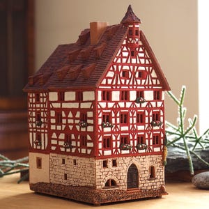 Può includere: Una casetta di pan di zenzero in miniatura in ceramica, di colore rosso e bianco. La casa ha un tetto a due falde e finestre decorative, e si trova su una base in pietra.
