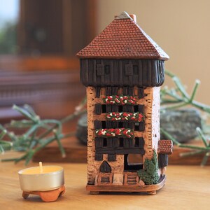 Op de afbeelding: Een klein, bruin keramiek huis met een rood dak en zwarte afwerking. Het huis heeft ramen en een deur, en er zijn bloemen op de balkons. Het huis staat op een houten oppervlak.