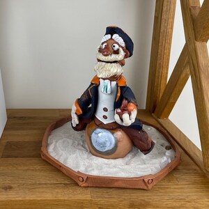 Può includere: Una statuetta in ceramica di un marinaio con la barba bianca e una giacca blu e arancione, seduto su un oggetto rotondo e marrone con sabbia bianca intorno. La statuetta è su uno scaffale di legno.
