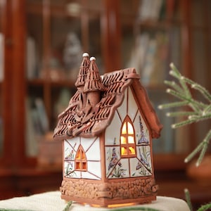 May include: A small, hand-crafted ceramic house with a white and brown glaze. The house has a detailed roof and windows with a light inside. The house is sitting on a hessian surface.