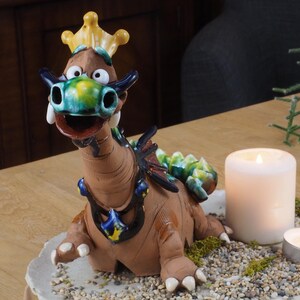 Op de afbeelding: Een keramiek drakenbeeldje met een kroon, groene en blauwe schubben en een wijd open mond. De draak zit op een wit bord met kleine witte stenen en groen mos. Er zijn twee kaarsen op het bord, een witte en een kleine zilveren.