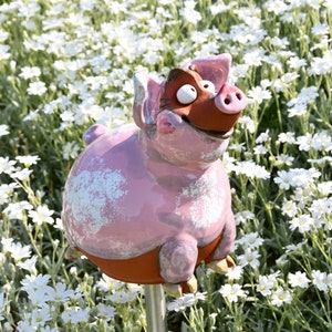Può includere: Una stravagante statua da giardino a forma di maiale rosa con dettagli marroni e un'espressione sorridente. Il maiale in ceramica è ambientato su uno sfondo di fiori bianchi e fogliame verde. La statua è montata su un paletto di metallo.