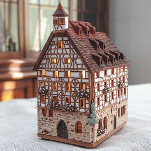 May include: A ceramic model of a traditional German half-timbered house with a red tiled roof, white walls, and brown beams. The house has multiple windows with glowing lights inside, and there are small flower boxes on the balconies. The house is sitting on a white tablecloth.