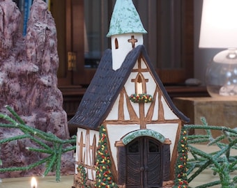 May include: A small, white ceramic church with a black roof and green trim. The church has a cross on the top and a wreath on the door. There is a lit candle in front of the church.