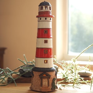 Op de afbeelding: Een keramisch vuurtorenbeeldje met rode en witte strepen en een klein huis aan de basis. De vuurtoren staat op een houten tafel met groene takken en stenen.