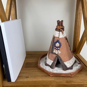 May include: A small ceramic teepee with brown, white and blue details sits on a wooden shelf. The teepee has a white base with a brown rim and a small opening at the bottom.