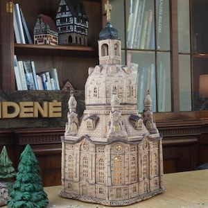 Puede incluir: Un modelo de cerámica detallado de una iglesia con una cúpula y una cruz en la parte superior. El modelo está iluminado desde el interior, creando un brillo cálido. El modelo está sentado en una mesa de madera con un letrero que dice "MIDENÉ".