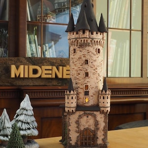 Op de afbeelding: Een keramisch torenmodel met een zwart dak en een lichtbruine bakstenen buitenkant. De toren heeft een grote boogdeur en verschillende kleine ramen. De toren is van binnenuit verlicht, waardoor een warme gloed ontstaat.