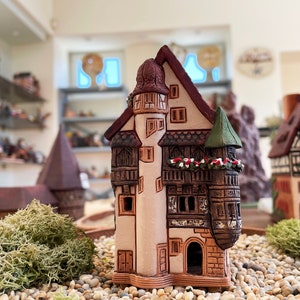 Può includere: Una casa in miniatura in ceramica con un tetto verde e una facciata marrone e bianca. La casa ha una torre con un tetto rosso e un piccolo balcone. La casa è circondata da piccole pietre e muschio verde.