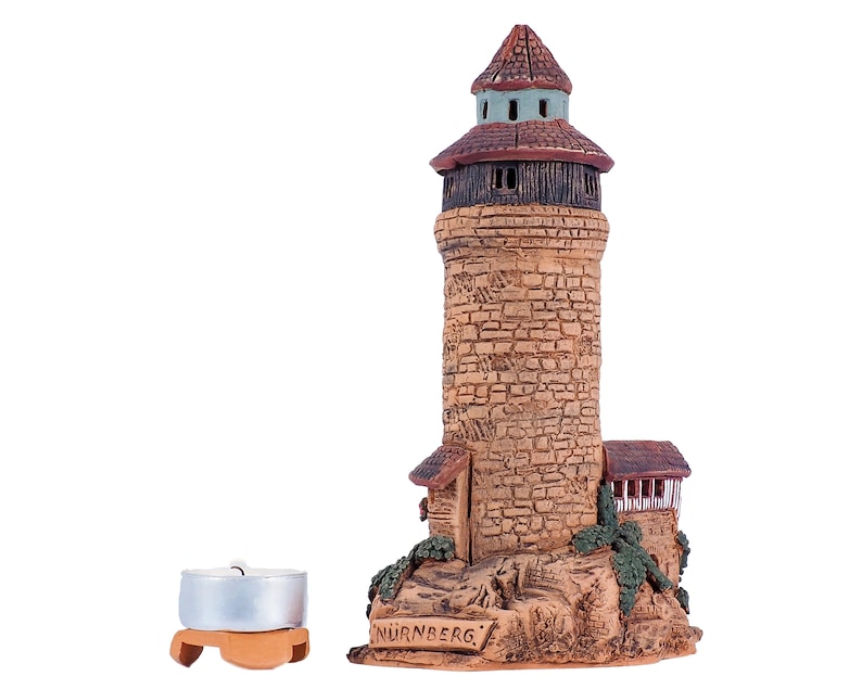 May include: A ceramic model of a tower with a brown roof and stone walls. The tower is labeled "NURNBERG". A small, silver candle holder is in the foreground.
