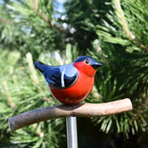 May include: A ceramic bird figurine with black, white, and red paint, perched on a brown branch. The bird is sitting on a metal rod.