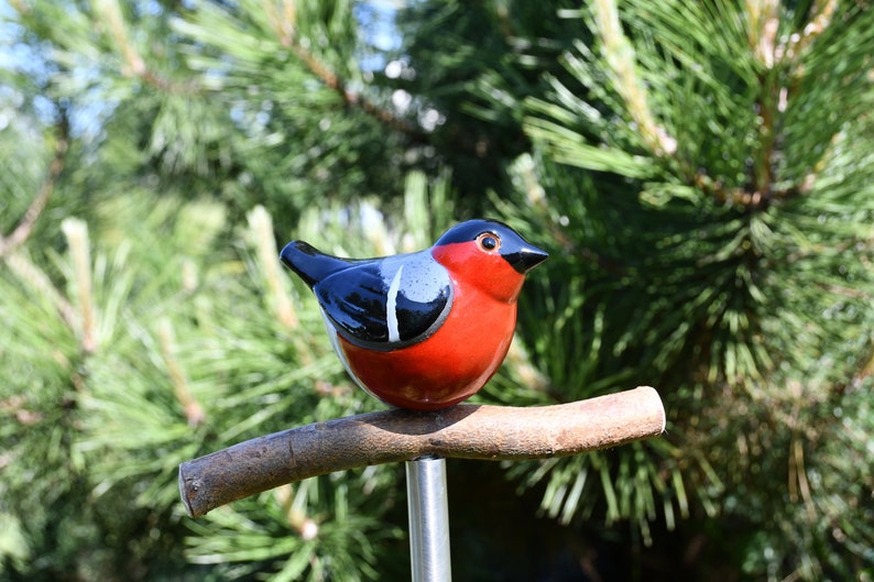May include: A ceramic bird figurine with black, white, and red paint, perched on a brown branch. The bird is sitting on a metal rod.