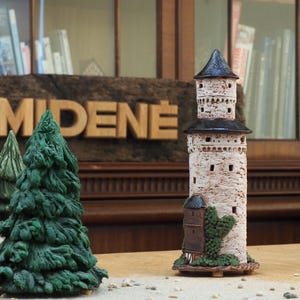 May include: A detailed ceramic model of a medieval tower with a black roof and small windows. The tower is light beige with a textured surface. Green ceramic trees and the wooden letters "MIDENE" are in the background.