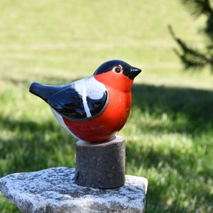 May include: Ceramic bird figurine with black, white, and red paint. The bird is perched on a small wooden stump.