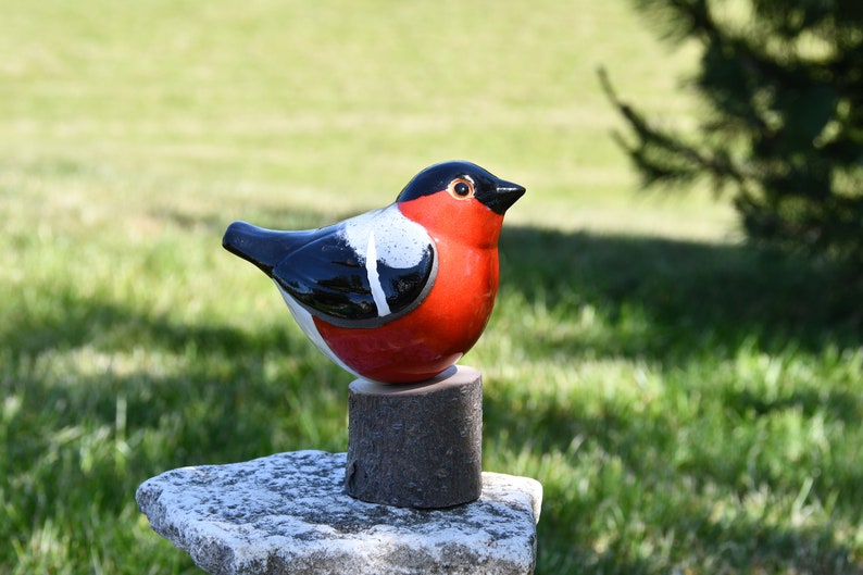May include: Ceramic bird figurine with black, white, and red paint. The bird is perched on a small wooden stump.