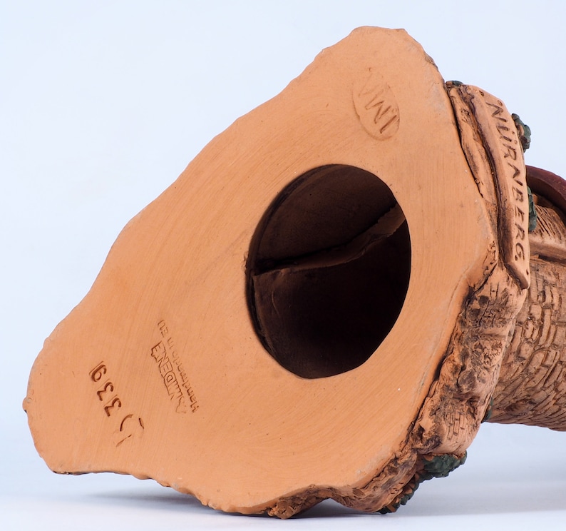 May include: A terracotta figurine of a house with a thatched roof and a chimney. The figurine is hand-painted with brown, green, and white details. The bottom of the figurine is stamped with "NURNBERG" and "INI" in a circle.