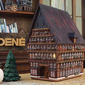 Puede incluir: Un modelo detallado de una casa tradicional alemana de entramado de madera con un techo marr&oacute;n, molduras blancas y un letrero que dice "MIDENE". La casa tiene varias ventanas y una peque&ntilde;a puerta. El modelo est&aacute; sentado en una mesa con una vela y dos peque&ntilde;os pinos.