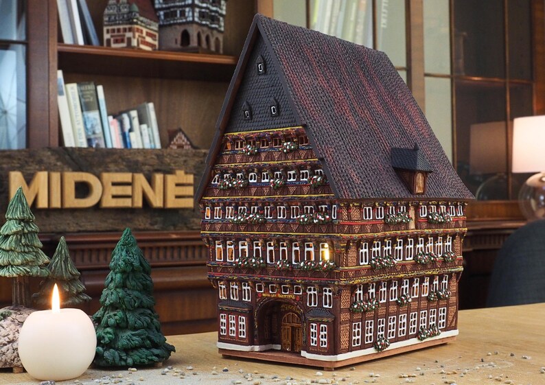 Puede incluir: Un modelo detallado de una casa tradicional alemana de entramado de madera con un techo marr&oacute;n, molduras blancas y un letrero que dice "MIDENE". La casa tiene varias ventanas y una peque&ntilde;a puerta. El modelo est&aacute; sentado en una mesa con una vela y dos peque&ntilde;os pinos.