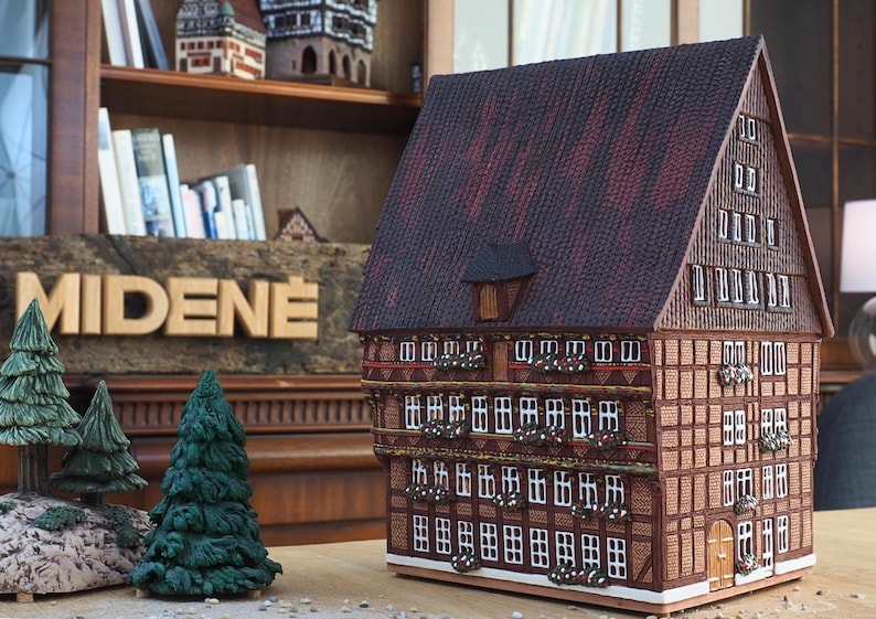 Puede incluir: Un modelo detallado de una casa tradicional alemana de entramado de madera con techo marr&oacute;n, ventanas blancas y vigas de madera decorativas. El modelo est&aacute; hecho de madera y tiene un letrero que dice "MIDENE".