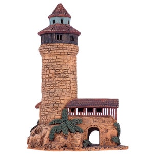 May include: A small, hand-painted ceramic tower with a brown roof and a stone-like exterior. The tower has a small arched doorway and a window with white trim. The tower is surrounded by green foliage.
