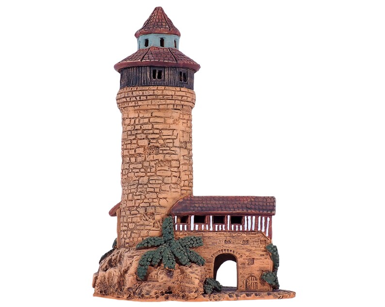 May include: A small, hand-painted ceramic tower with a brown roof and a stone-like exterior. The tower has a small arched doorway and a window with white trim. The tower is surrounded by green foliage.