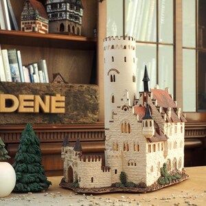 Könnte beinhalten: Ein detailliertes Modell einer mittelalterlichen Burg mit einem hohen Turm, aufwendigen Steinmetzarbeiten und einem roten Ziegeldach. Das Modell ist aus weißen und braunen Materialien gefertigt und hat kleine Fenster und Türen. Die Burg steht auf einem Holztisch mit einem Schild, auf dem "MIDENE" steht.