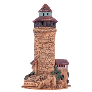 May include: A ceramic model of a tower with a brown roof and a green top. The tower is made of brown and tan bricks and has a small window on each side. The base of the tower is made of brown and tan rocks and has the word "NURNBERG" written on it.