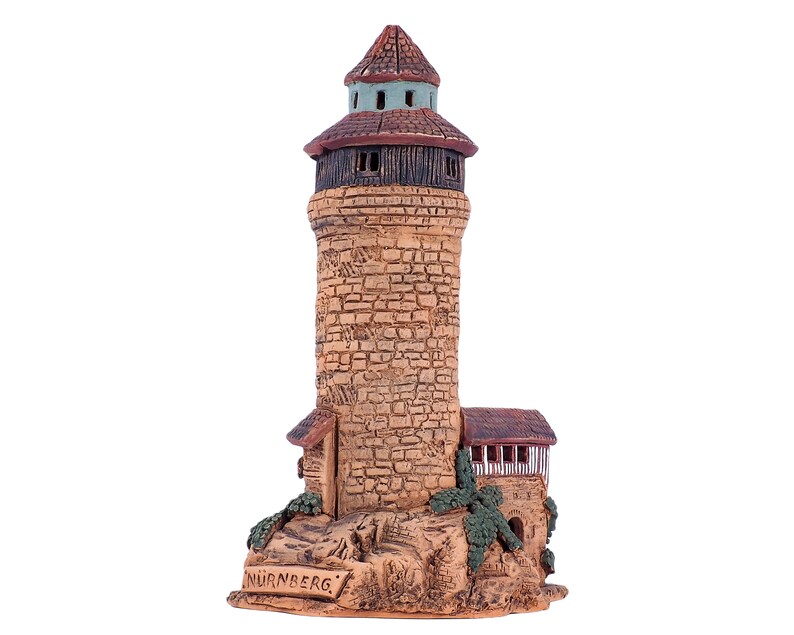 May include: A ceramic model of a tower with a brown roof and a green top. The tower is made of brown and tan bricks and has a small window on each side. The base of the tower is made of brown and tan rocks and has the word "NURNBERG" written on it.