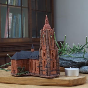 Peut inclure: Une figurine détaillée d'église en terre cuite avec un haut clocher et un toit sombre. Le bâtiment miniature est posé sur une base en bois, à côté d'une bougie chauffe-plat allumée. L'arrière-plan comprend une armoire en bois et de la verdure.