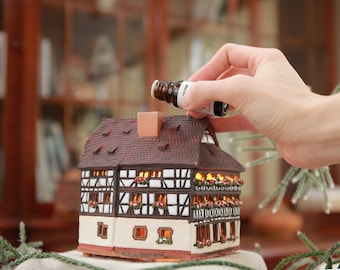 May include: A white ceramic house with a brown roof and black trim. The house has windows with lights inside and a chimney. A hand is holding a small brown bottle with a label that says "Winter"