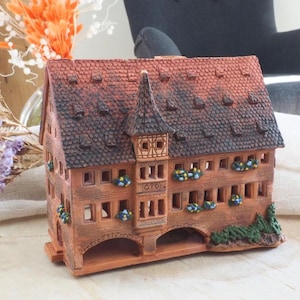May include: A detailed ceramic model of a European-style building with a red tile roof and a tower. The building has multiple windows with small floral decorations and is painted in shades of brown and red.