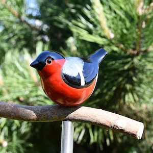 May include: A ceramic bird figurine with black, red, and white paint, perched on a brown branch. The bird is sitting on a metal pole.