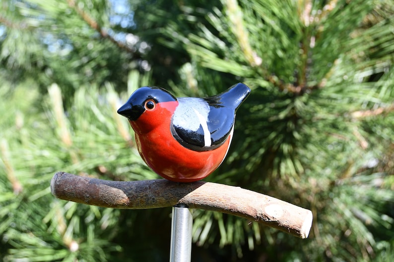 May include: A ceramic bird figurine with black, red, and white paint, perched on a brown branch. The bird is sitting on a metal pole.
