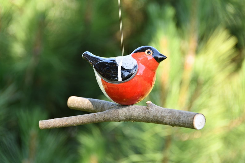 May include: A ceramic bird ornament with black, white, and red paint, perched on a brown branch. The bird is hanging from a string.