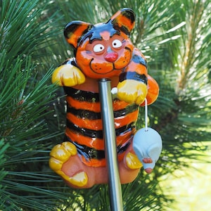 以下が含まれることがあります： オレンジと黒の縞模様のセラミック製の虎の置物で、小さな青いネズミの置物を抱えています。虎は金属製の棒の上にいます。