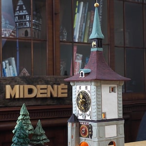 May include: A detailed ceramic model of a clock tower with a white face and a red roof. The tower is lit from within, and the clock face has Roman numerals. The model is sitting on a wooden table with a sign that reads "MIDENE".